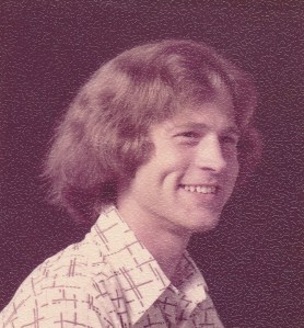 A faded color photo of a man in his twenties with longish hair. He is turning toward the viewer's right and smiling and wears a collared shirt.