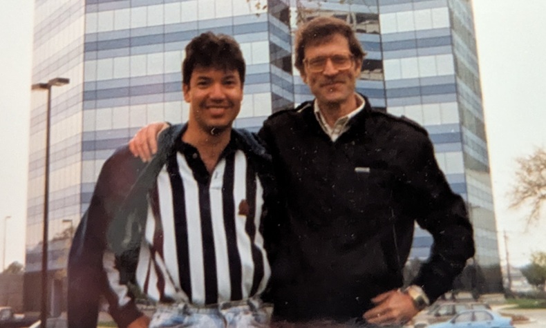 A Mexican-American man in a shirt with white and black vertical stripes, and a white man with glasses and a dark jacket, who has his arm around the other. They are standing in the parking lot of a tall office building.